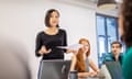 A woman stands in a meeting room, presenting to peers, holding a piece of paper.