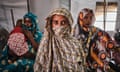A woman with a covered head and mouth looks directly at the camera. Behind her another woman sits on a bed comforting a small child, while another woman sits beside her, looking towards the camera.