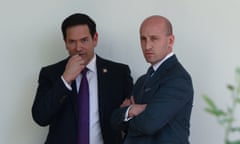 Two men in suits stand and talk in front of a white wall outside