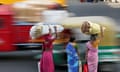 Three women in saris with goods balanced on their head waiting to cross a road as a truck and tuktuk speed past them