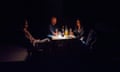 Three people sit with drinks at a table lit by a battery lamp amid darkness