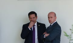 Two men in suits stand and talk in front of a white wall outside
