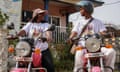 Two women wearing T-shirts that read 'Bike Ladies' sit on motorbikes looking at each other. One wears a helmet, the other a sun visor