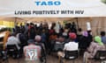 People sit facing away from the camera on chairs in a large tent labelled 'Taso' and 'Living positively with HIV'