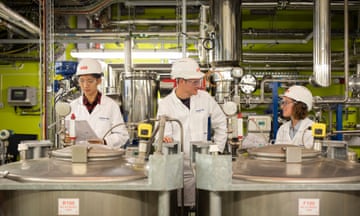 People in hard hats and lab coats inside a lab surrounded by large metal drums and other pieces of equipment