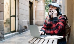 An older man laughs as he uses a phone and laptop while sitting at a table in an alleyway in Barcelona
