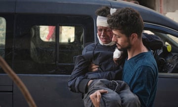 A boy grimacing in pain with a bandage on his head being carried into hospital by a man