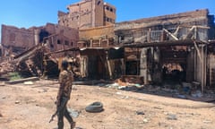 A man wearing camouflage patterned clothes and holding an automatic rifle walks past a burnt-out building.